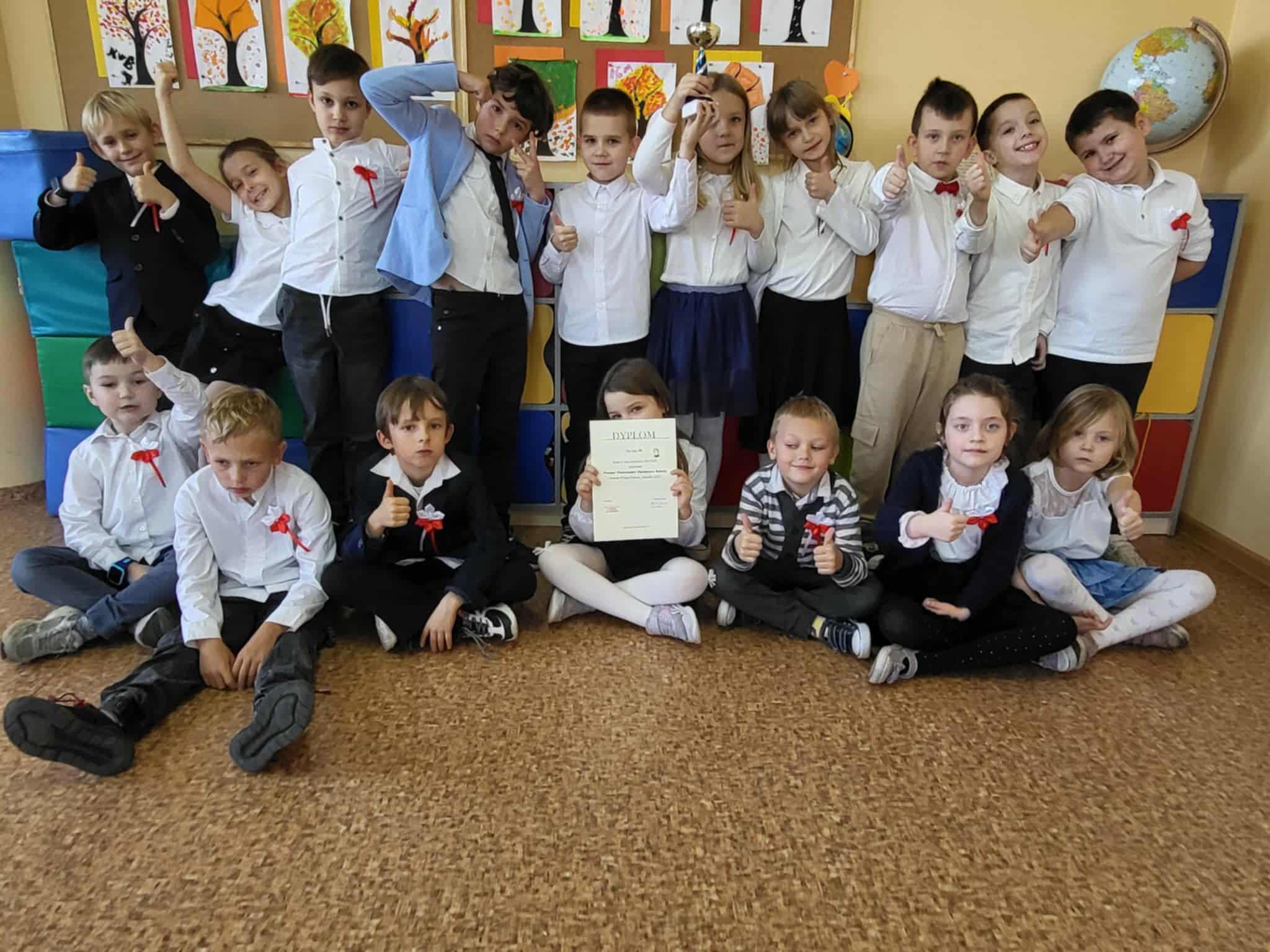 Uczniowie klasy 1b z SP 13 w Pabianicach pozują z dyplomem i pucharem za zwycięstwo w Stefankach; dzieci odświętnie ubrane, siedzą i stoją, pokazując kciuki do g&oacute;ry.