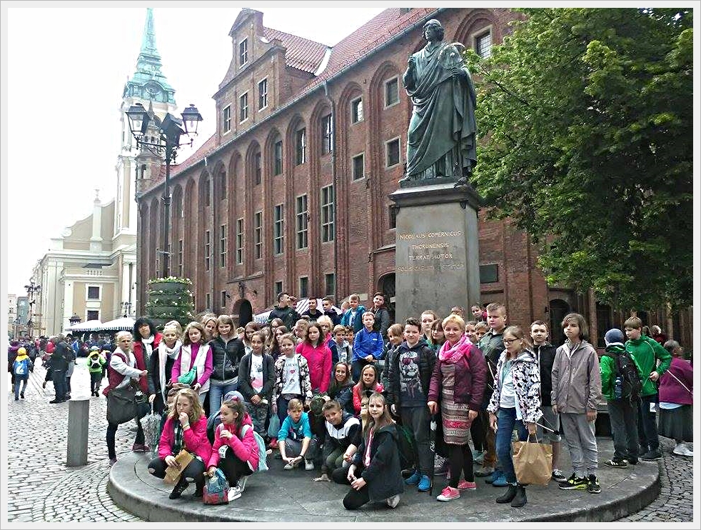 Wycieczka ucznió klas młodszych do Torunia. Grupa dzieci i opiekunów pozuje do zdjęcia grupowego przed pomnikiem Mikołaja Kopernika w Toruniu. Pomnik znajduje się na wysokim cokole z inskrypcją łacińską. W tle widoczna jest ceglana fasada Ratusza Staromiejskiego z arkadowymi oknami oraz wieża kościoła św. Ducha. Na placu widać brukowaną nawierzchnię, drzewa oraz tłumy turystów. Zdjęcie zrobione podczas wycieczki szkolnej.