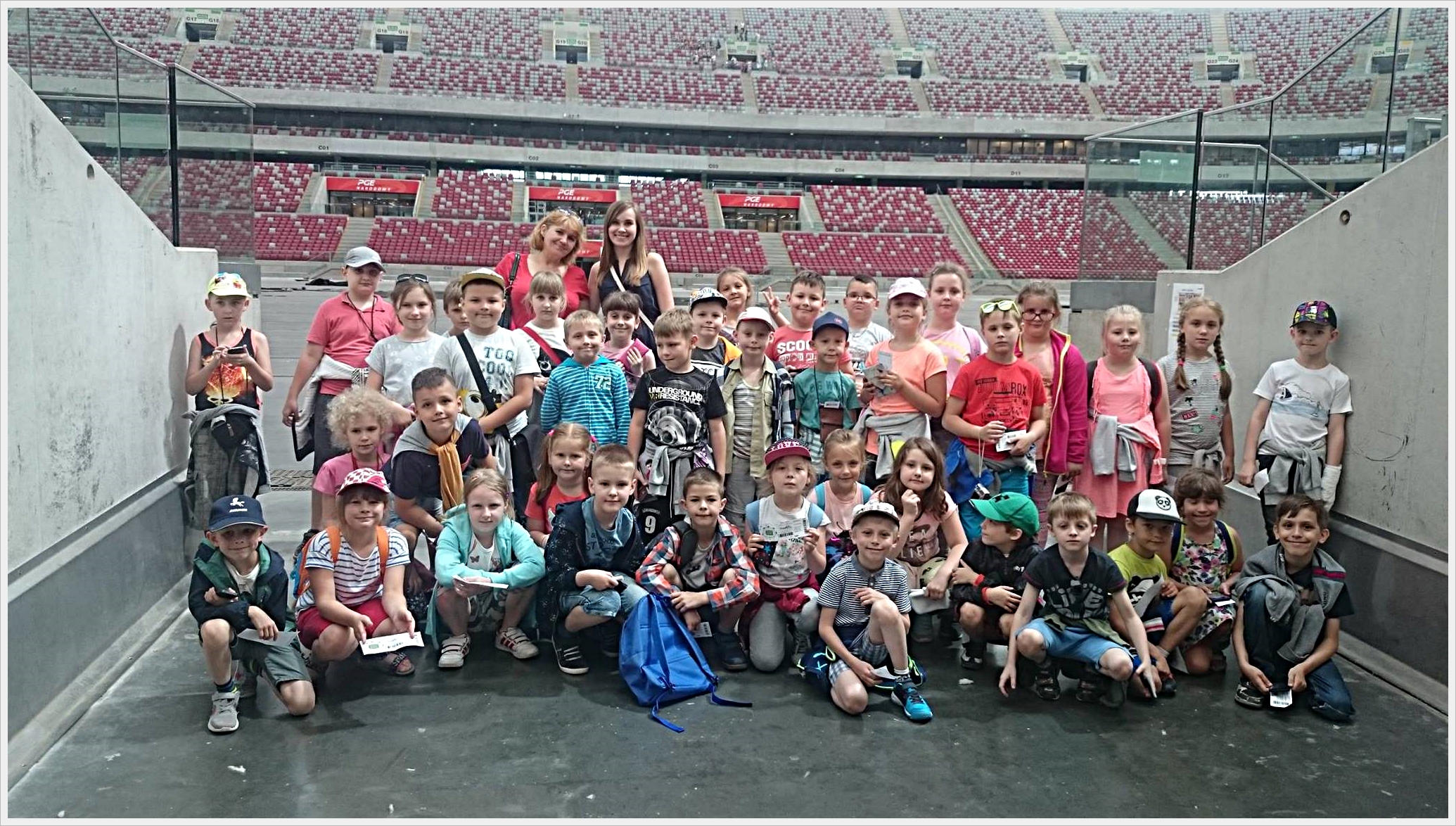 Wycieczka klas trzecich do Warszawy Duża grupa dzieci pozuje do wspólnego zdjęcia na tle stadionu z czerwono-białymi miejscami na trybunach. Dzieci stoją i kucają między betonowymi ścianami w tunelu prowadzącym na murawę. W tle widać ogrom trybun, co sugeruje, że zdjęcie wykonano na dużym stadionie sportowym, prawdopodobnie podczas wycieczki szkolnej. Dzieci są ubrane letnio, wiele z nich ma czapki z daszkiem, plecaki i bilety w rękach. Towarzyszą im dwie uśmiechnięte opiekunki.