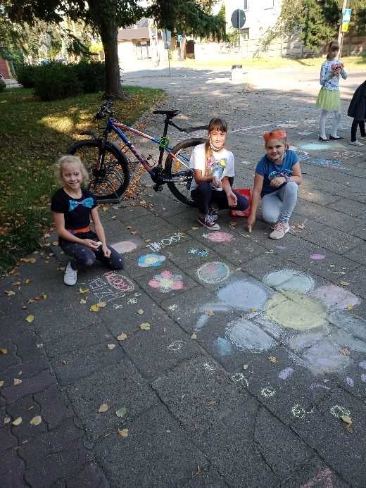 Niecodzienna lekcja. Trzy dziewczynki siedzą na chodniku i uśmiechają się do zdjęcia, otoczone kolorowymi rysunkami wykonanymi kredą. Na chodniku widać m.in. rysunek dużego kwiatka i różne kształty w intensywnych kolorach. W tle stoi rower, a po prawej stronie widać inne dzieci oraz drzewa rzucające cień na chodnik.