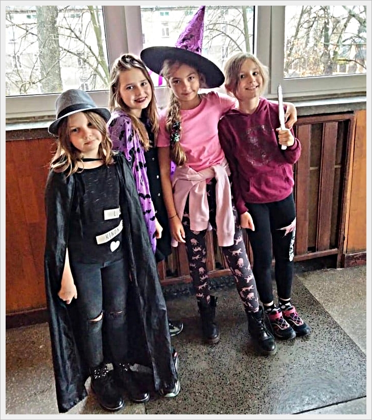 Ostatki w naszej szkole. Cztery dziewczynki stoją obok siebie w szkolnym korytarzu, przebrane w kolorowe stroje, prawdopodobnie z okazji Halloween lub balu przebierańców. Pierwsza z lewej ma na sobie czarne ubranie z peleryną, kapelusz i napisy na koszulce. Druga dziewczynka nosi fioletową pelerynę w czarne nietoperze. Trzecia jest ubrana w różową bluzkę, legginsy z różowymi kotami i ma na głowie duży kapelusz czarownicy. Czwarta dziewczynka ubrana jest w bordową bluzę i trzyma w ręku różdżkę. Wszystkie stoją w uśmiechniętej pozie na tle okien z widokiem na drzewa i budynki za nimi.