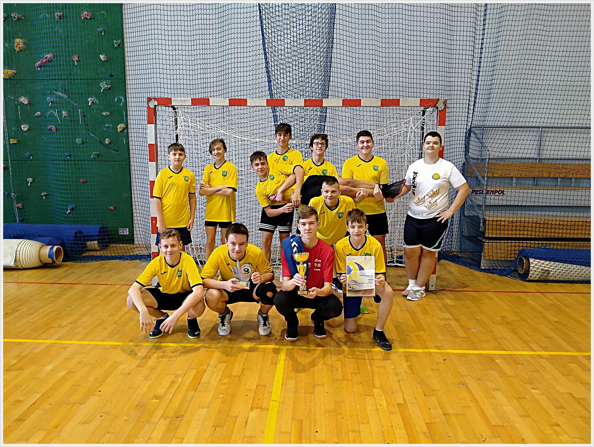 Zawody sporotwe - piłka ręczna. Grupa chłopców w żółtych koszulkach sportowych pozuje w sali gimnastycznej na tle bramki do piłki ręcznej. Trzech z nich klęczy z przodu, trzymając puchar i dyplom. W tle widać ściankę wspinaczkową, siatki zabezpieczające i zrolowane maty gimnastyczne. Wszyscy są uśmiechnięci i wyglądają na zadowolonych z osiągnięcia sportowego.