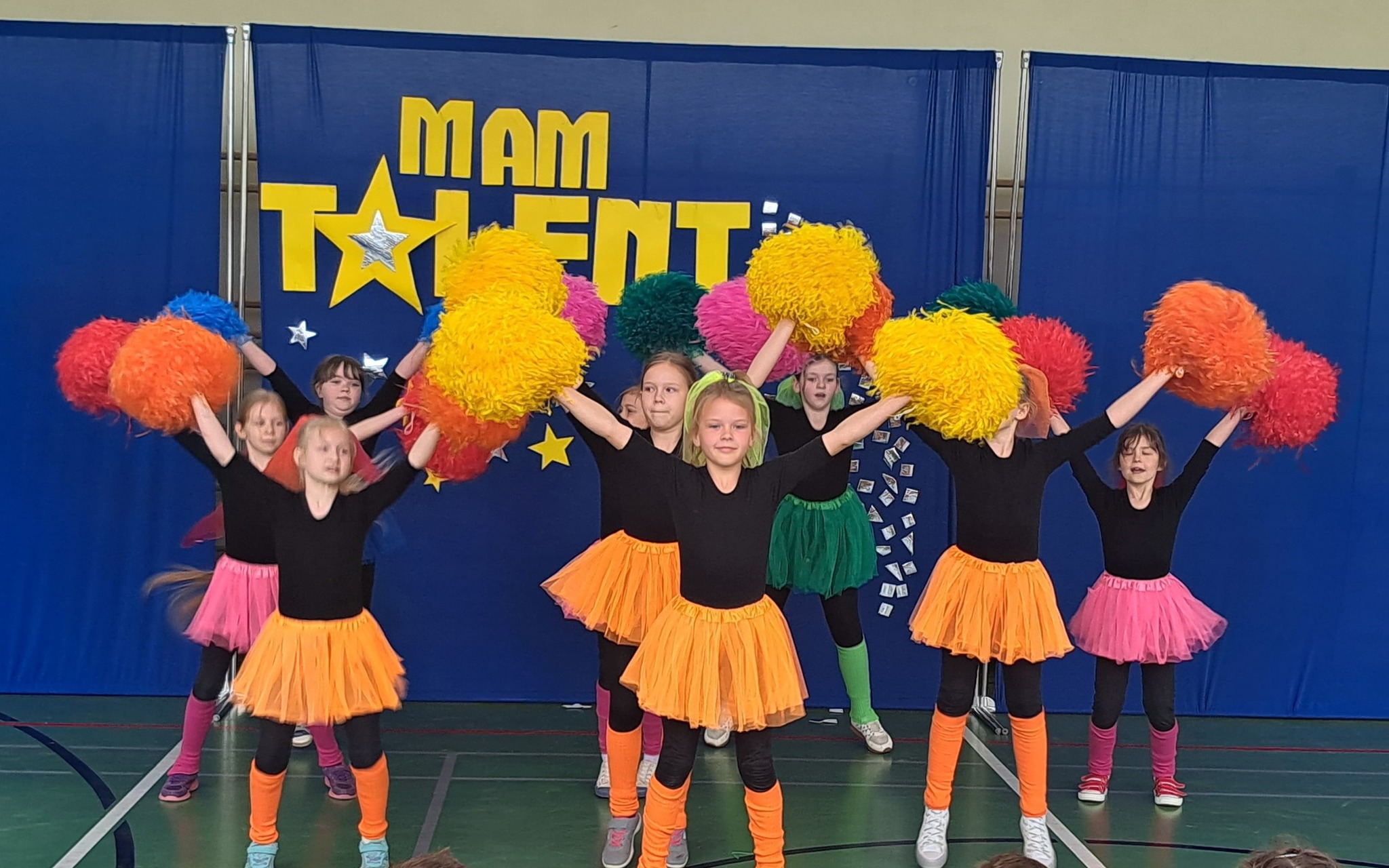 Pokaz "Mam talent" Grupa dziewczynek występuje na scenie podczas szkolnej edycji programu „Mam Talent”. Ubrane są w czarne koszulki, kolorowe tiulowe spódniczki oraz legginsy i opaski w intensywnych barwach. Każda z nich trzyma kolorowe pompony w dłoniach. W tle widać niebieski parawan z żółtym napisem „MAM TALENT” oraz dekoracjami w postaci gwiazdek.