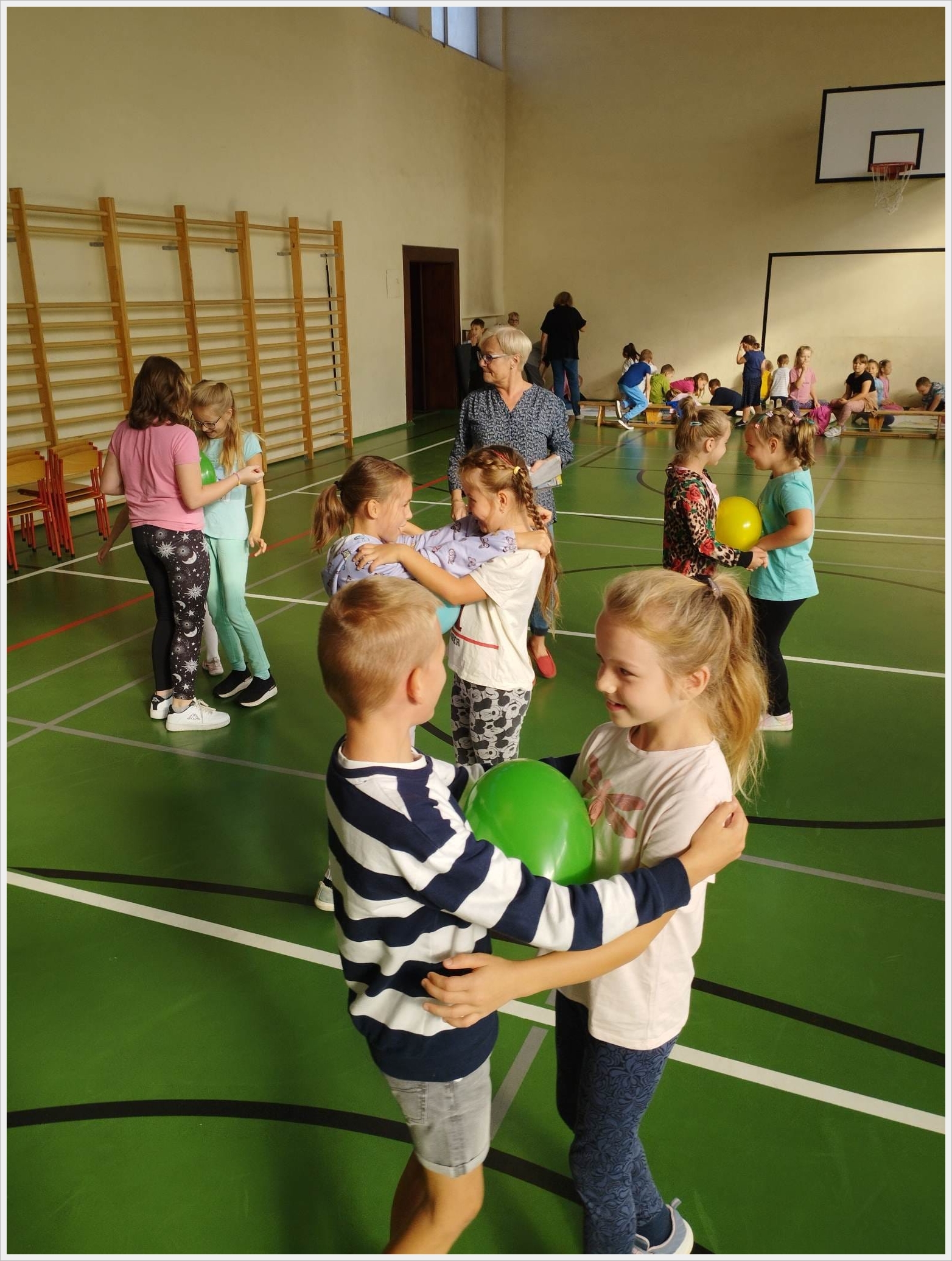Zajęcia sportowe świetliczaków. Dzieci biorą udział w zabawie ruchowej w sali gimnastycznej. W parach trzymają balony pomiędzy swoimi ciałami, ćwicząc współpracę i równowagę. W tle widoczne są inne grupy dzieci, nauczyciele oraz sprzęt sportowy. Na ścianie znajduje się drabinka gimnastyczna i tablica do koszykówki. Atmosfera jest radosna i aktywna.