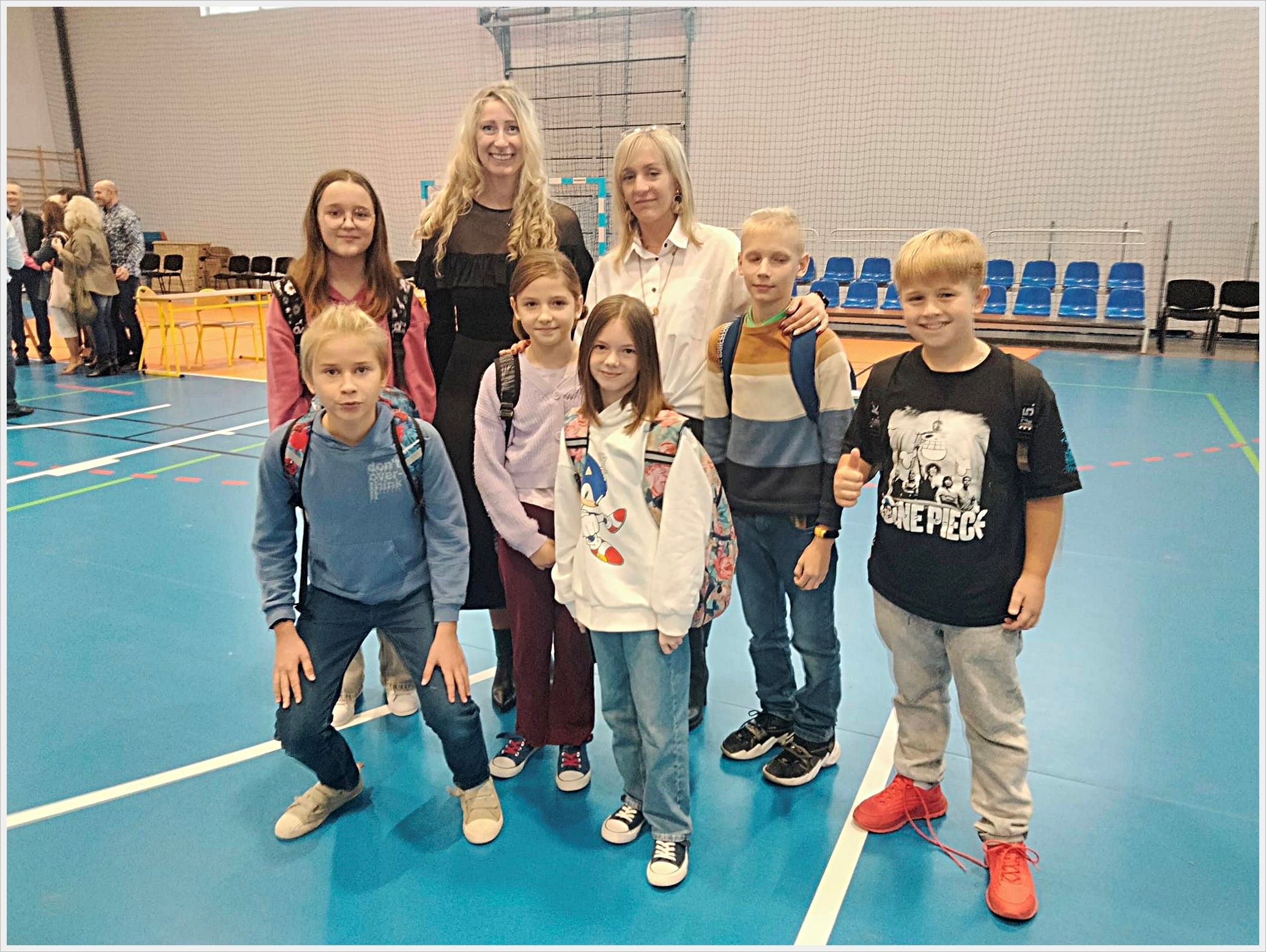 Spotkanie z Pabianickimi olimpijczykami. Grupa siedmiorga uczniów i olimpijki Aleksandry Shelton (Socha) wraz z nauczycieleką pozuje do zdjęcia na sali gimnastycznej. Dzieci mają na sobie codzienne ubrania i plecaki, a jedna z dziewczynek nosi bluzę z postacią Sonica. Aleksandra Shelton (Socha) i nauczycielki stoją z tyłu i uśmiechają się. W tle widać niebieskie trybuny, siatkę na ścianie oraz inne osoby rozmawiające przy stolikach.