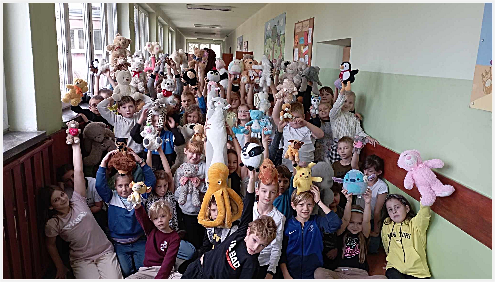 Dzień pluszaka. Grupa dzieci siedzi i stoi w szkolnym korytarzu, trzymając w górze swoje pluszowe maskotki. Dzieci są uśmiechnięte i radosne, a maskotki są różnorodne – od misiów, przez pingwiny i koty, po ośmiornice i postacie z bajek. Scena odbywa się w jasnym korytarzu z dużymi oknami po lewej stronie. W tle widać kolorowe plakaty na ścianach.