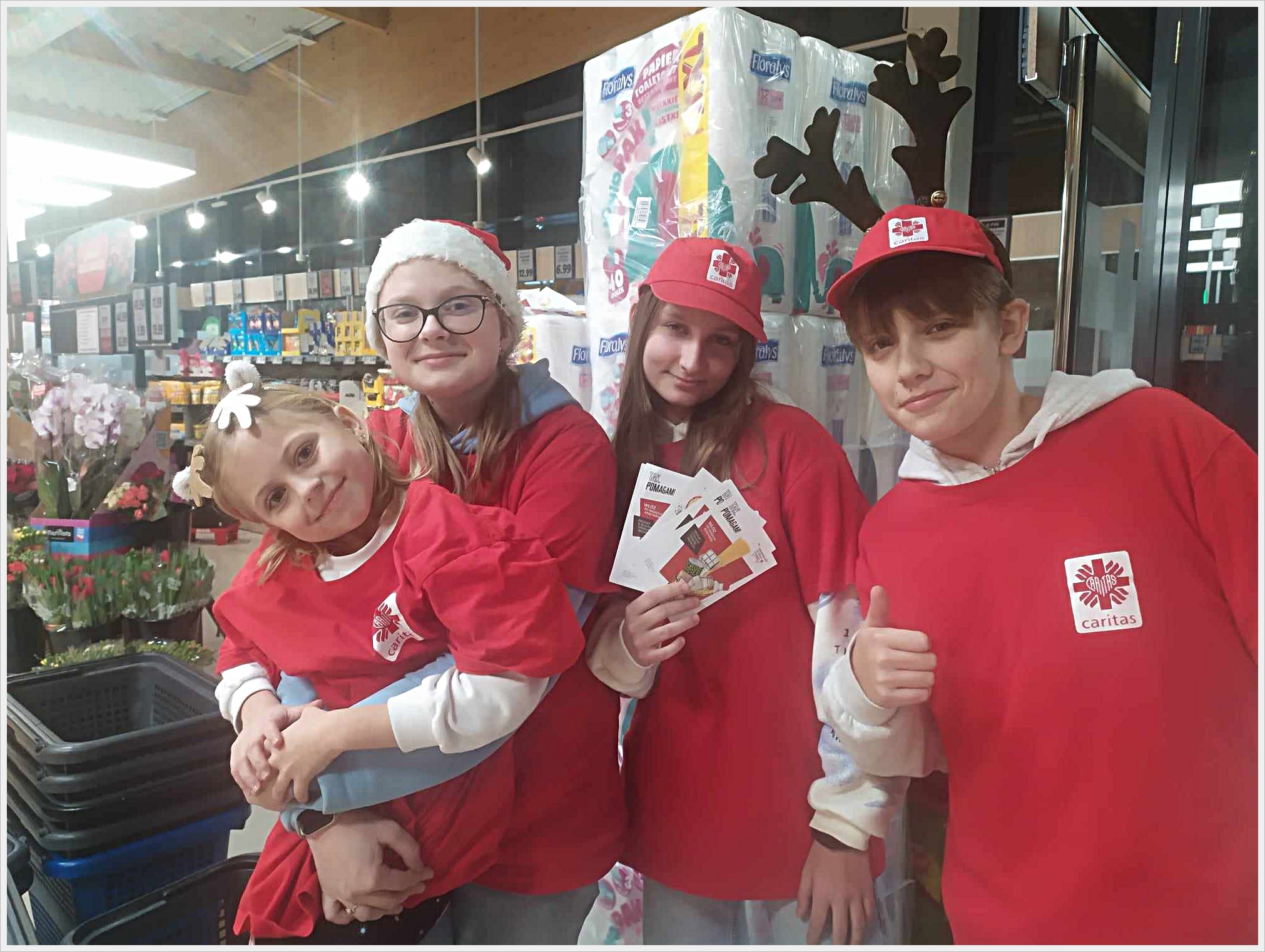 Wolontariusz naszej szkoły. Czworo dzieci ubranych w czerwone koszulki z logo Caritas pozuje do zdjęcia w sklepie spożywczym. Jedna dziewczynka trzyma na rękach młodszą dziewczynkę z opaską w kształcie kwiatka. Druga dziewczynka trzyma w rękach ulotki, a chłopiec po prawej stronie pokazuje gest „ok” z uniesionym kciukiem. Dzieci mają na głowach świąteczne akcenty – czapki Mikołaja i opaskę z rogami renifera. W tle widać kwiaty, wózki sklepowe i regały z produktami.