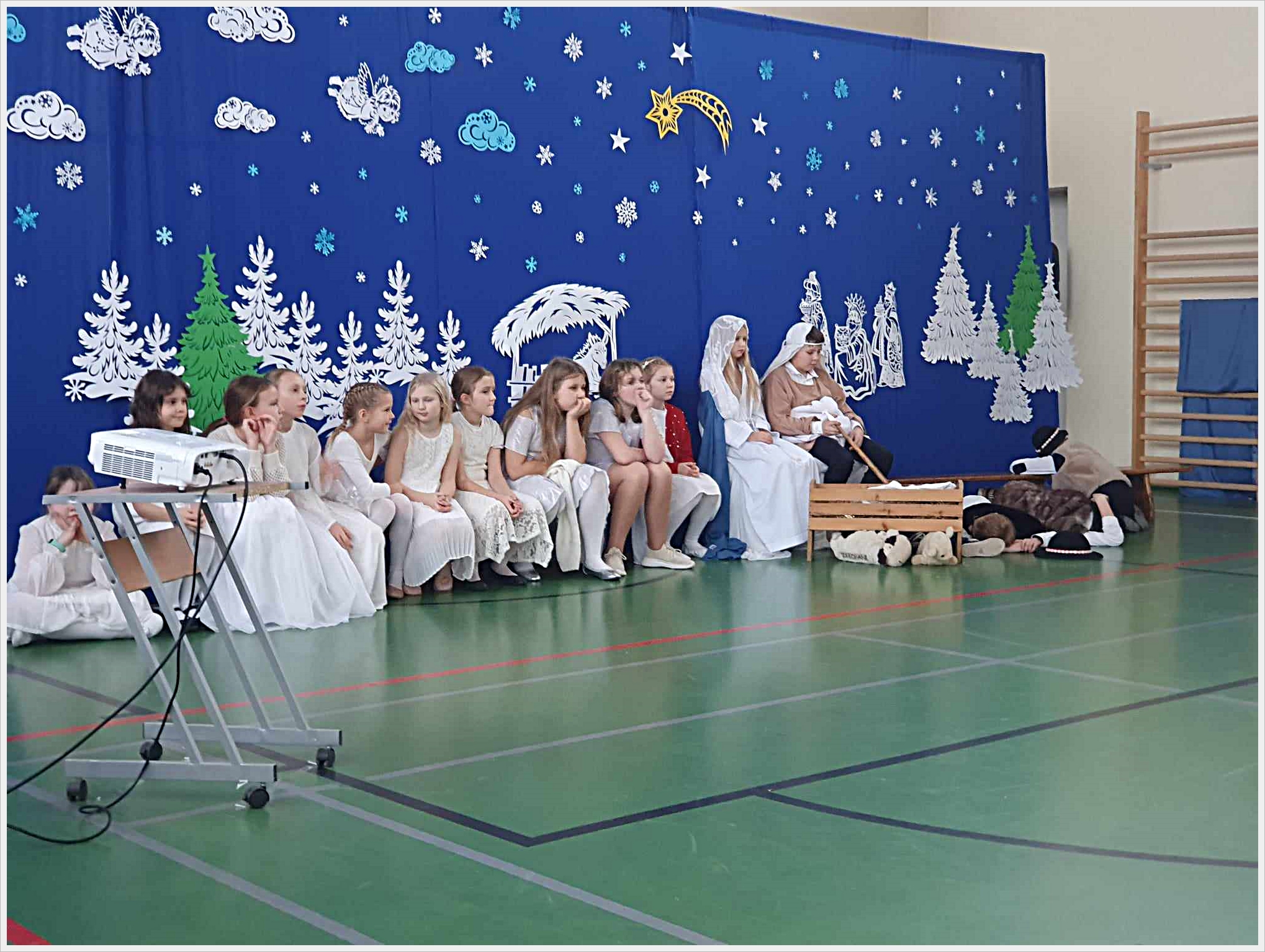 Jasełka szkolne. Grupa dzieci bierze udział w przedstawieniu jasełkowym w sali gimnastycznej. Na tle niebieskiej dekoracji z motywami zimowymi (gwiazdy, płatki śniegu, chmurki, aniołki, choinki) dzieci siedzą na ławce lub na podłodze. Po prawej stronie scena z Maryją, Józefem i lalką w roli Dzieciątka Jezus w drewnianym żłóbku, otoczona przez dzieci przebrane za pasterzy i zwierzęta. Przed grupą stoi projektor na metalowym stoliku. Całość utrzymana w świątecznym, zimowym klimacie.