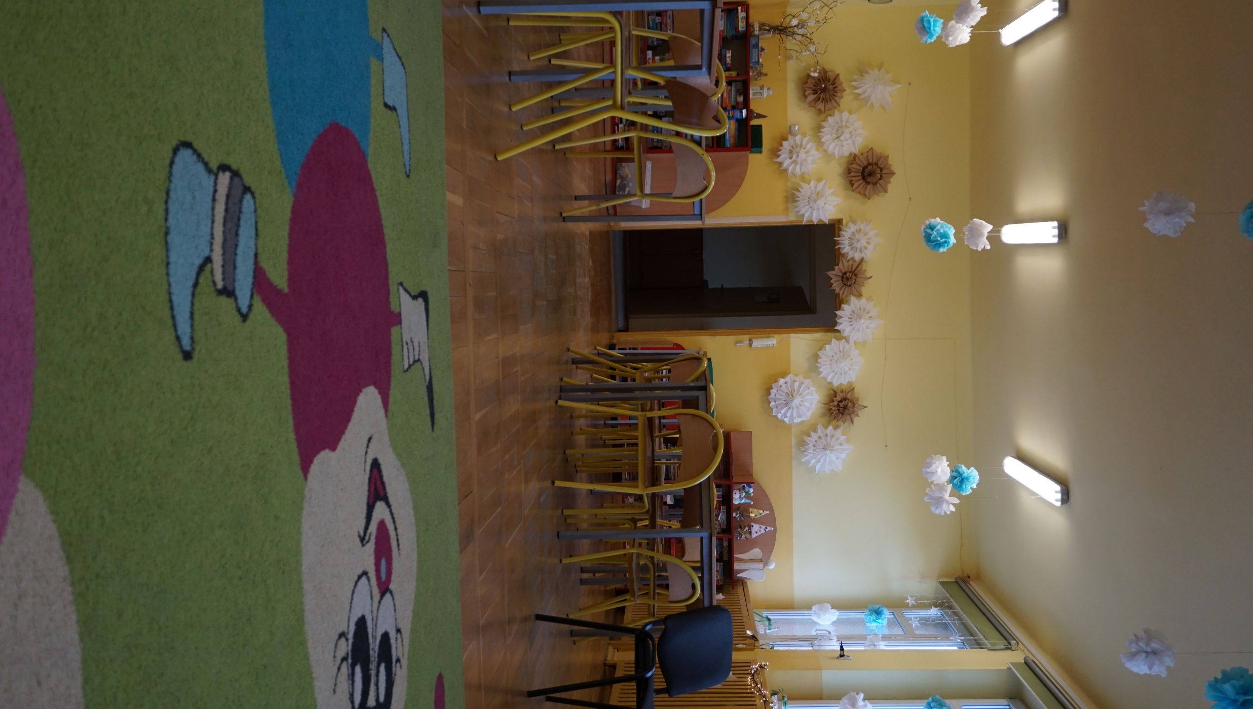 Świetlica szkolna. Sala z jasnożółtymi ścianami i drewnianą podłogą. Na pierwszym planie widoczny jest kolorowy dywan z rysunkiem uśmiechniętej postaci z dużymi okrągłymi segmentami ciała. W tle znajdują się dziecięce stoliki i krzesła oraz regały z grami i zabawkami. Na ścianie nad drzwiami zawieszone są dekoracje przypominające papierowe śnieżynki w kolorach białym i brązowym. Z sufitu zwisają białe i niebieskie pompony. Po prawej stronie – okna z ozdobami i lampkami.