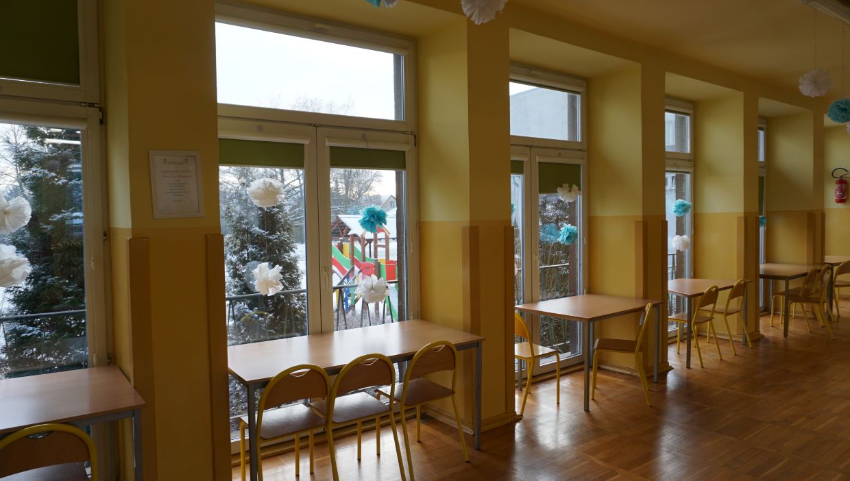 Świetlica szkolna. Jasna, żółta sala z dużymi oknami i drzwiami balkonowymi wychodzącymi na plac zabaw z kolorowymi zjeżdżalniami, częściowo zasłoniętymi śnieżnymi drzewami. Przy każdym oknie ustawione są proste drewniane stoliki z krzesłami. Na szybach zawieszone są papierowe dekoracje w kształcie białych i niebieskich pomponów, nadające wnętrzu zimowy klimat. Na ścianie widoczny jest certyfikat lub dyplom, a w tle – gaśnica. Podłoga z drewnianych paneli, całość sprawia wrażenie stołówki lub świetlicy w placówce edukacyjnej.