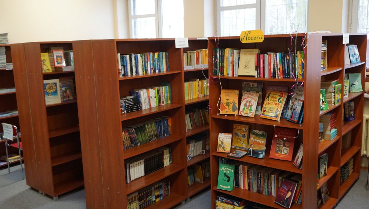 Biblioteka szkolna. Wnętrze szkolnej biblioteki z kilkoma drewnianymi regałami wypełnionymi kolorowymi książkami. Na jednym z regałów znajduje się żółty szyld z napisem „Nowości” oraz ozdobne serpentyny. Książki są ułożone pionowo i frontem do przodu – wśród nich widoczne są bajki, encyklopedie, książki przygodowe i edukacyjne. W tle jasne ściany i duże okna wpuszczające naturalne światło.
