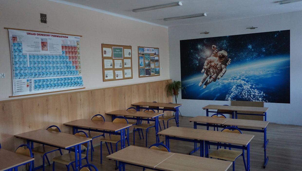 Sala fizyczna. Sala lekcyjna z ustawionymi w rzędach ławkami i krzesłami. Na ścianie po lewej stronie wisi kolorowy układ okresowy pierwiastków oraz tablice informacyjne. Na ścianie z tyłu znajduje się duża fototapeta przedstawiająca astronautę w przestrzeni kosmicznej nad Ziemią.