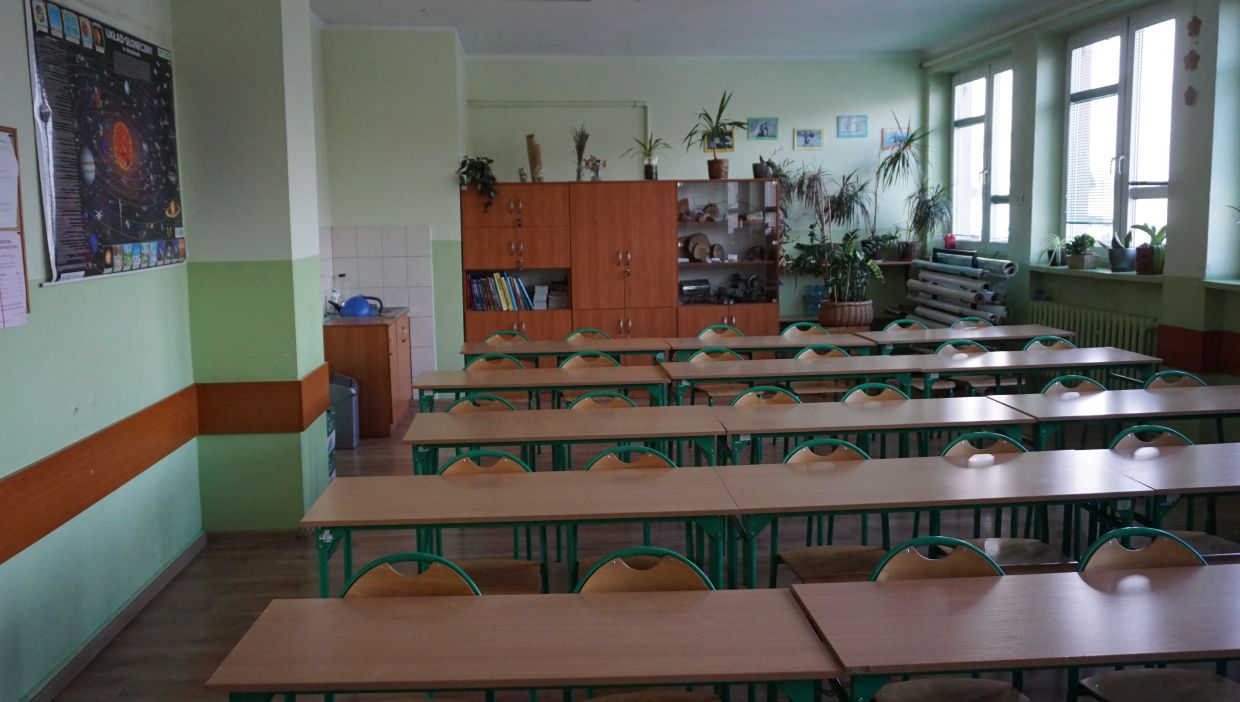Sala geograficzna. Sala lekcyjna z rzędami pustych ławek i krzeseł. Po lewej stronie widoczny plakat przedstawiający Układ Słoneczny. Z tyłu klasy znajduje się szafka z książkami, eksponatami i licznymi roślinami doniczkowymi. Pod oknami po prawej stronie również ustawiono wiele roślin.