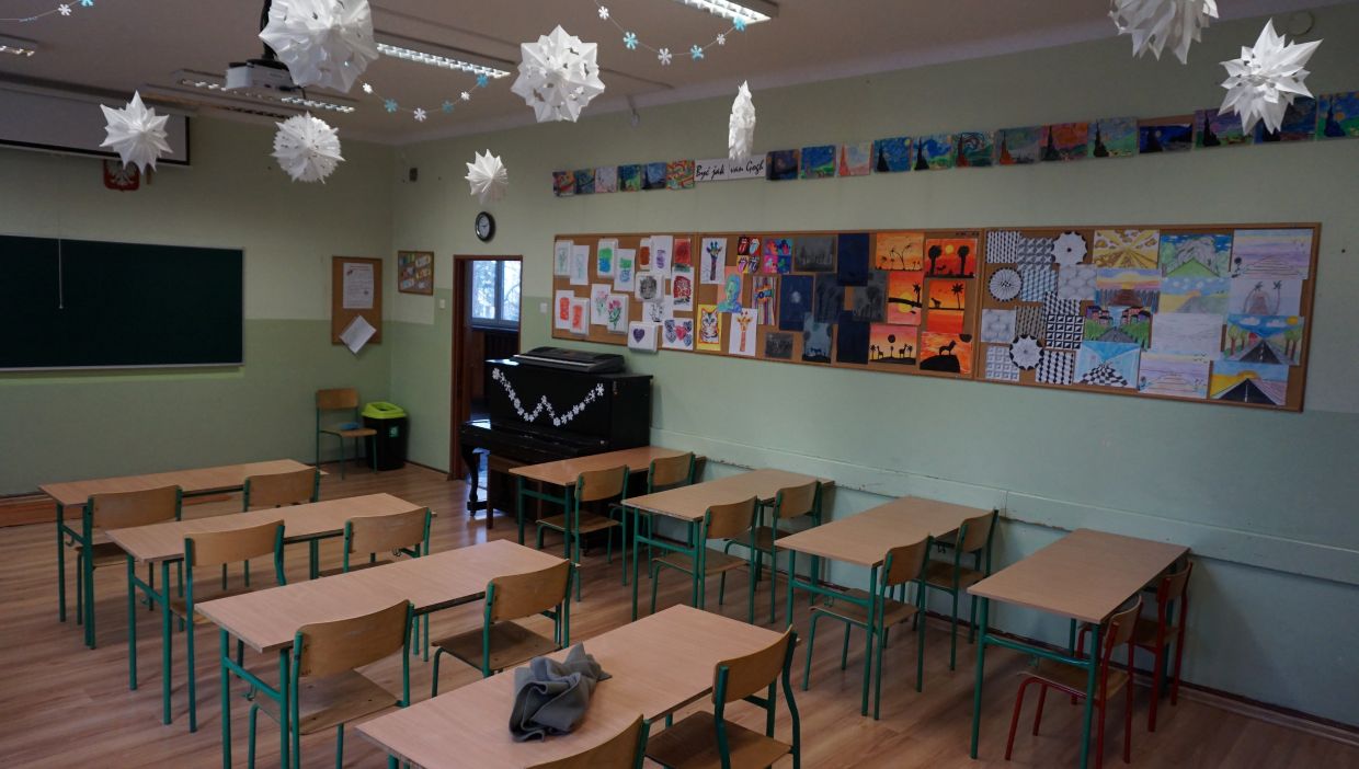 Sala języka polskiego. Sala lekcyjna z pustymi ławkami i zielonymi krzesłami. Na ścianie z prawej strony znajdują się tablice korkowe z kolorowymi rysunkami uczniów. Nad tablicami wiszą dodatkowe prace plastyczne. Z sufitu zwisają papierowe ozdoby w kształcie śnieżynek. W rogu klasy stoi czarny pianino ozdobione śnieżynkami.