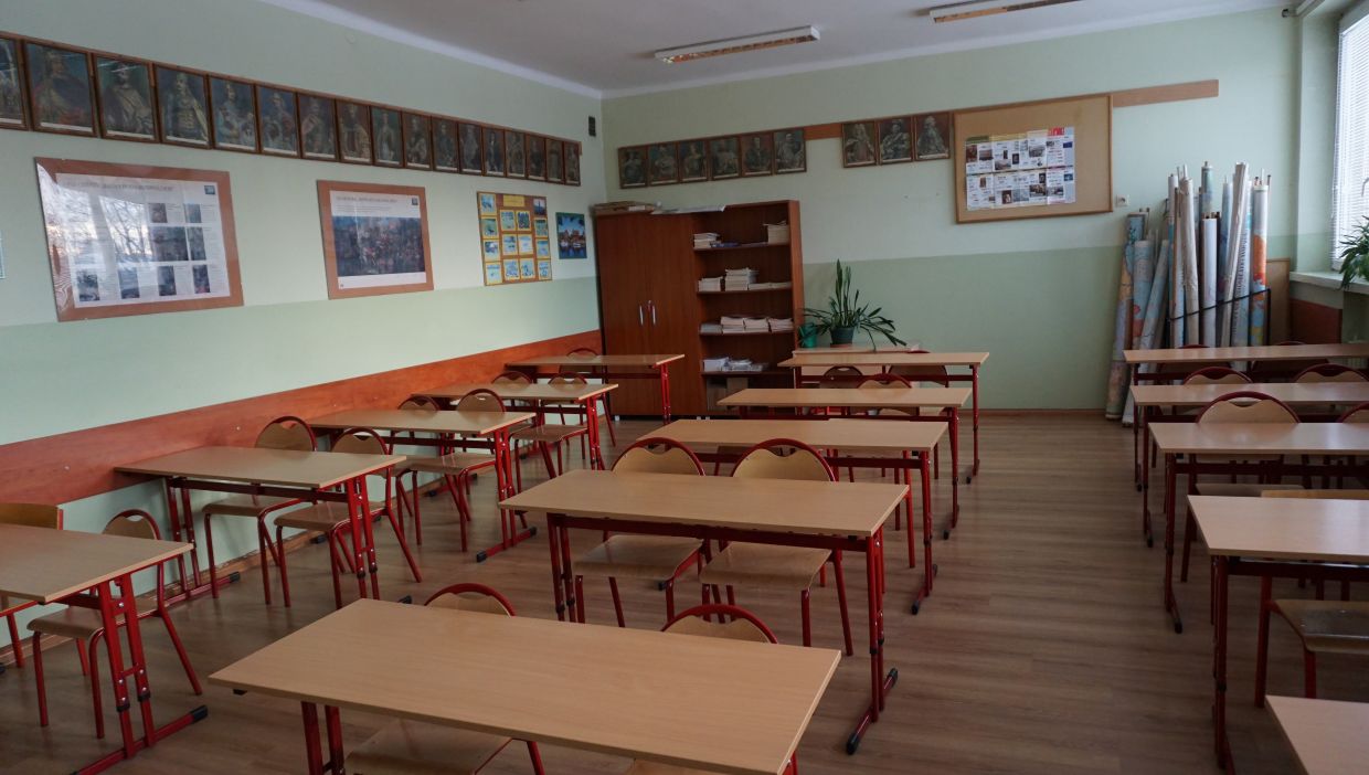 Sala historyczna. Sala lekcyjna w szkole z jasnymi, zielono-białymi ścianami. W pomieszczeniu znajdują się rzędy dwuosobowych ławek i krzeseł z czerwonymi metalowymi stelażami oraz jasnobrązowymi blatami i oparciami. Po lewej stronie widać plansze edukacyjne i obrazy historyczne w ramach, zawieszone w dwóch rzędach na ścianie. Z tyłu klasy znajduje się drewniana szafa oraz regał z ułożonymi stosami książek. Po prawej stronie, przy oknie, stoją zwinięte mapy w tubach oraz doniczki z roślinami. Podłoga pokryta jest panelami imitującymi drewno, a przez okno wpada dzienne światło.