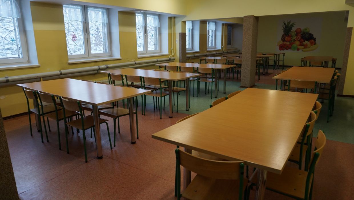 Stołówka. Szkolna stołówka z jasnymi żółtymi ścianami i wieloma prostokątnymi stołami otoczonymi drewnianymi krzesłami z metalowymi, zielonymi nogami. W pomieszczeniu jest kilka rzędów okien z firankami, przez które widać śnieg i gałęzie drzew. Na tylnej ścianie widnieje duża ilustracja przedstawiająca kompozycję owoców, m.in. ananasa, banana i winogron. Pod oknami biegną poziome rury grzewcze, a podłoga składa się z kolorowych segmentów w odcieniach czerwonym i zielonym.