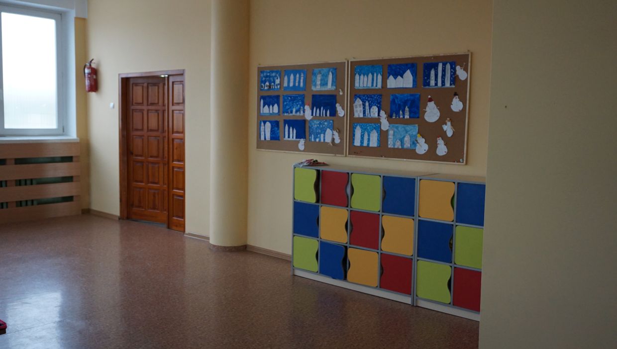 Sala klasy pierwszej. Sala z żółtymi ścianami, na których znajdują się dwie tablice korkowe z pracami plastycznymi przedstawiającymi zimowe krajobrazy z domkami i bałwankami. Poniżej znajduje się rząd kolorowych szafek uczniowskich w kolorach: czerwonym, żółtym, zielonym i niebieskim. Po lewej stronie widoczne są drewniane drzwi oraz okno z parapetem i grzejnikiem osłoniętym drewnianą osłoną.