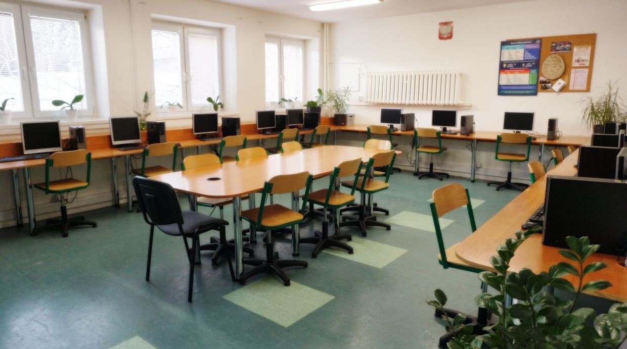 Sala informatyczna. Sala komputerowa w szkole z jasnymi ścianami i zieloną wykładziną podłogową. Wzdłuż ścian ustawione są biurka z komputerami stacjonarnymi oraz krzesła z zielonymi oparciami. Na środku pomieszczenia znajduje się duży, owalny stół z krzesłami do pracy grupowej. Pomieszczenie jest dobrze oświetlone naturalnym światłem wpadającym przez trzy okna, na których parapetach stoją doniczki z roślinami. Na ścianie wisi tablica korkowa z informacjami oraz plakat edukacyjny. W pomieszczeniu znajduje się także kilka większych roślin w donicach.