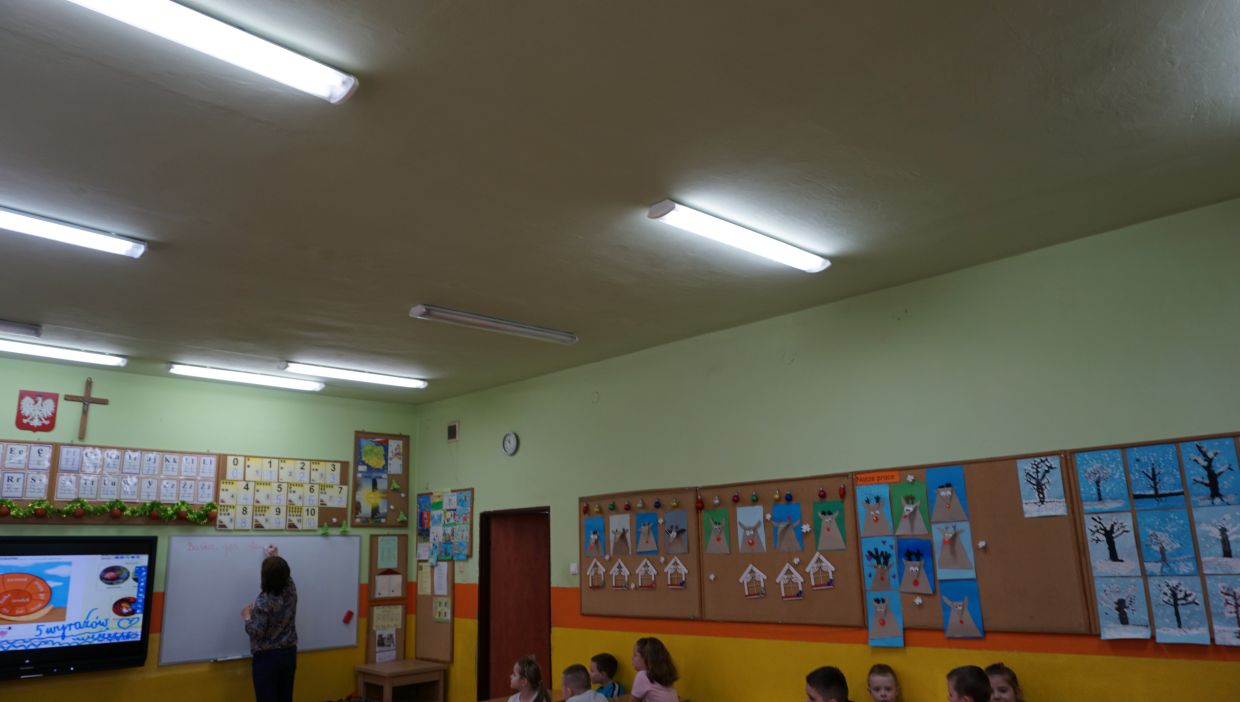 Sala klasy trzeciej. Sala lekcyjna z jasnozielonymi ścianami, w której nauczycielka pisze na białej tablicy, a grupa uczniów siedzi na krzesełkach z boku. Na ścianach wiszą plansze edukacyjne, kolorowe prace plastyczne dzieci oraz tablice z cyframi i literami. Widoczne są także krzyż i godło Polski.