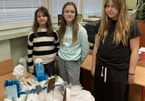 Trzy uczennice stoją przy makiecie zimowej Narnii z rzeką, drzewami i elementami zabudowy.