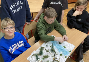 Uczniowie klasy piątej prezentują makietę zimowej Narnii z rzeką i ośnieżonym krajobrazem inspirowaną lekturą C. S. Lewisa.