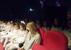 Uczennice klasy 2b w białych strojach aniołów siedzą na widowni przed występem podczas akcji Aniołkowe Granie.