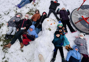 Dzieci leżą na śniegu wokół bałwana, tworząc śnieżne anioły.