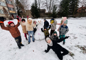 Grupa dzieci stoi wokół bałwana ulepionego ze śniegu na szkolnym boisku.