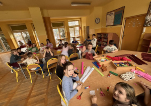 Dzieci siedzą przy stolikach i wykonują prace z kolorowej bibuły. Na stołach widoczne są nożyczki, klej i materiały plastyczne. W tle znajduje się regulamin świetlicy i dekoracje zimowe.