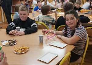 Uczniowie siedzą przy stołach w klasie i jedzą wspólną kolację.