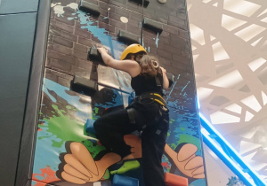 Uczennica wspina się po ściance wspinaczkowej w kasku i uprzęży.