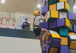 Osoba wspina się po kolorowej konstrukcji wspinaczkowej w uprzęży.