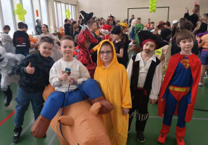 Grupa dzieci w przebraniach karnawałowych stoi w sali gimnastycznej podczas balu.