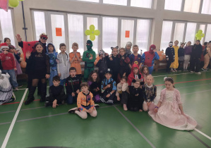 Liczna grupa dzieci w kostiumach karnawałowych siedzi i stoi w sali gimnastycznej podczas wspólnej fotografii.