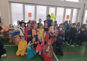 Dzieci w kolorowych przebraniach stoją w sali gimnastycznej podczas wspólnego zdjęcia.