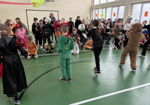 Grupa dzieci w strojach karnawałowych bierze udział w zabawie z piłkami w sali gimnastycznej.