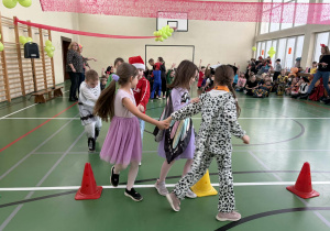Dzieci w przebraniach trzymają się za ręce i przechodzą między pachołkami podczas zabawy ruchowej w sali gimnastycznej.