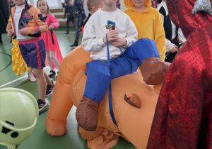 Chłopiec w kostiumie z nadmuchiwanym koniem stoi w sali gimnastycznej wśród innych przebranych uczniów.