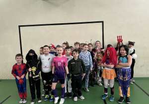 Grupa dzieci w różnorodnych strojach karnawałowych stoi w sali gimnastycznej i pozuje do wspólnego zdjęcia.