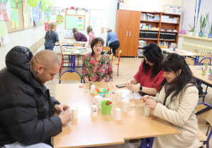 Grupa dorosłych siedzi przy stole i wykonuje prace plastyczne z papierowych kubków.