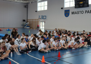 Uczniowie siedzą na podłodze hali sportowej i słuchają podsumowania wydarzenia.