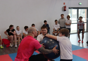 Damian Janikowski instruuje dwóch uczniów podczas ćwiczenia z piłką gimnastyczną w hali sportowej.
