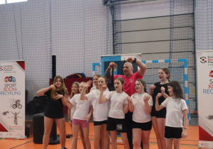 Damian Janikowski stoi z grupą uczennic w hali sportowej na tle baneru „Sport Kocha Recykling”.
