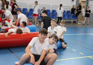 Grupa uczniów uczestniczy w sportowej konkurencji z użyciem dmuchanych materacy w hali sportowej.