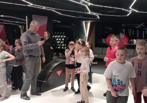 Dzieci stoją przy torze do kręgli, a w tle rodzice obserwują zabawę w kręgielni.