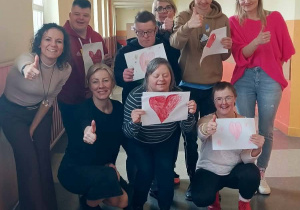 Grupa dorosłych i uczniów trzyma rysunki serc i uśmiecha się do zdjęcia na szkolnym korytarzu.