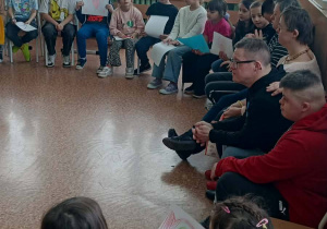 Dzieci i goście siedzą w kręgu w klasie, trzymają rysunki serc i uczestniczą w zajęciach integracyjnych.