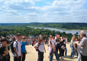 rupa młodzieży uczestniczy w wycieczce na wzniesieniu z panoramicznym widokiem na rzekę, lasy i miasteczko z czerwonymi dachami. Uczniowie stoją na piaszczystym podłożu, słuchając przewodnika, który opowiada i gestykuluje. Dzieci noszą plecaki i sportowe ubrania, część osłania oczy przed słońcem. Na dalszym planie widać inne grupy turystów oraz malowniczy krajobraz pod niebem z białymi chmurami.