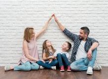Czteroosobowa rodzina siedzi na podłodze przy białej ceglanej ścianie. Rodzice, siedząc po bokach, unoszą dłonie i łączą je nad głowami dzieci, tworząc symboliczny „dach”.