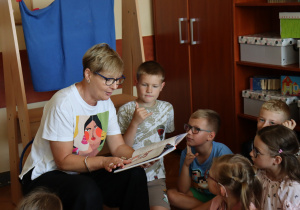 Nauczycielka Monika Metller siedzi w kręgu dzieci i czyta im książkę. Uczniowie uważnie słuchają, siedząc na podłodze w klasie. W tle znajdują się regały z materiałami dydaktycznymi i pudełkami.